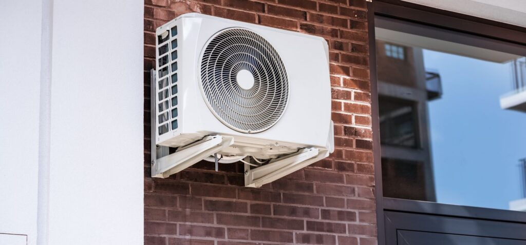 Wall-mounted outdoor mini-split HVAC unit installed on a brick exterior wall beside a window in a residential home.